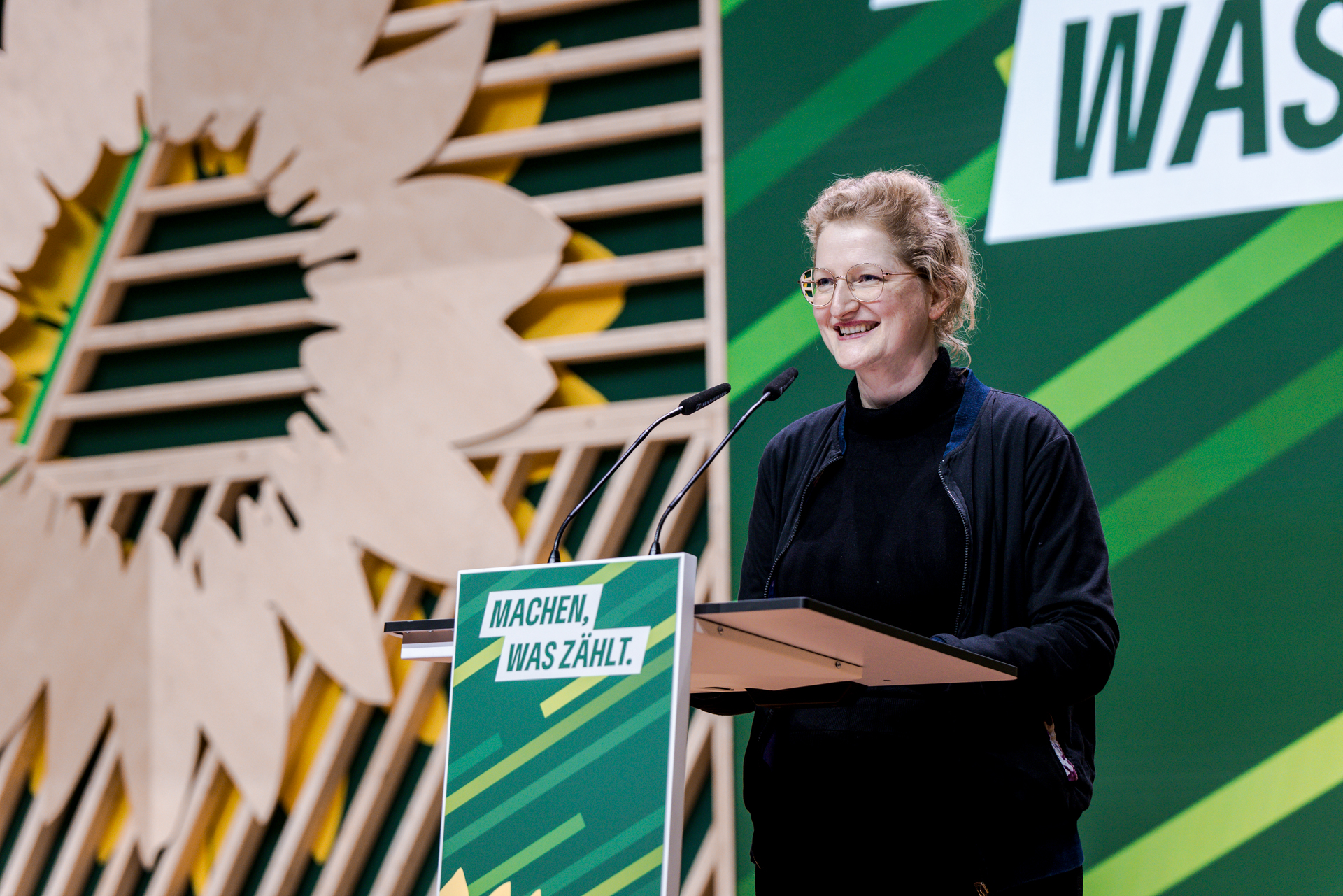 Andrea Wörle vor einem Redepult mit der Aufschrift "Machen, was zählt", Bühnenaufbau im Hintergrund mit großem Sonnenblumen-Logo, Holzleisten und Streifen in verschiedenen Grüntönen